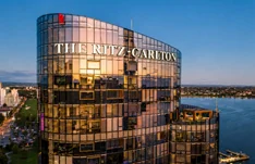 A curved glass hotel tower carrying the Ritz-Carlton name, lit at sunset above the waterfront